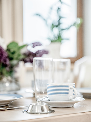 A white cup with blue stripes on a saucer sits on a table near a blurred vase of flowers and two tall glasses. The background is soft-focused, suggesting a bright and airy setting.