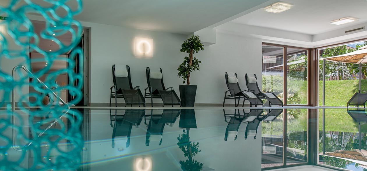 Indoor pool area with calm water reflecting lounge chairs and a potted plant. Glass doors open to a garden with grass and umbrellas, allowing natural light. A contemporary light fixture is on the wall to the left. A decorative element is in the foreground.