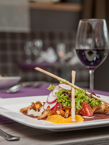 Ein Gourmetsalat mit Kirschtomaten, Radieschenscheiben, Blattgemüse und Grissini ist kunstvoll auf einem weißen Teller angerichtet. Im Hintergrund steht ein Glas Rotwein. Das Ambiente ist raffiniert, mit einer violetten Tischdecke und elegantem Besteck.