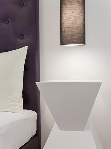 Minimalist bedroom with a geometric white nightstand, a purple tufted headboard, and crisp white bedding. A modern black lamp hangs above the nightstand, casting soft light. The decor is simple and elegant.