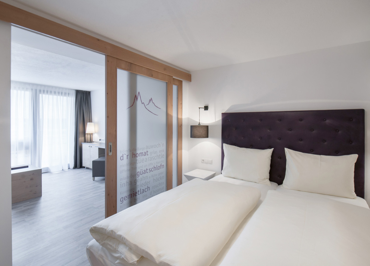 Modern hotel room with a large bed, dark upholstered headboard, and white bedding. A frosted glass sliding door with mountain design and German words partially separates the bedroom from a well-lit sitting area. Minimalist decor.