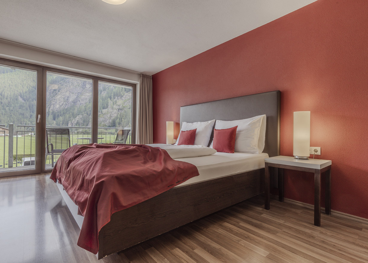 A modern hotel room with a large bed featuring a red blanket and pillows. Two bedside tables with lamps flank the bed. The wall behind the bed is red, and a large window provides a view of a green landscape and mountains.