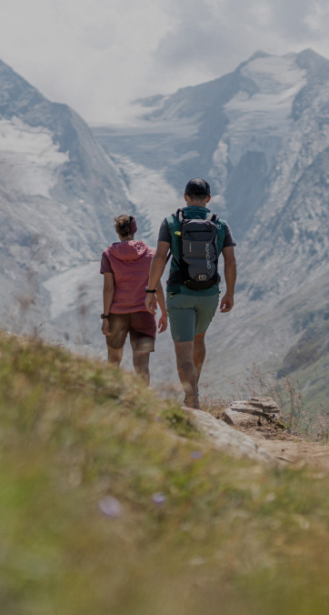 A couple hiking up a mountain trail with the man carrying a backpack. They are walking towards towering, snow-capped peaks under a cloudy sky. Wildflowers and grass surround the path, suggesting a late spring or summer setting.