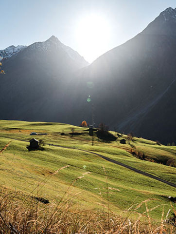 Sunlight illuminates a lush, green valley with a few small houses scattered among the fields. Majestic mountains rise in the background under a clear blue sky. Tall grass is visible in the foreground.