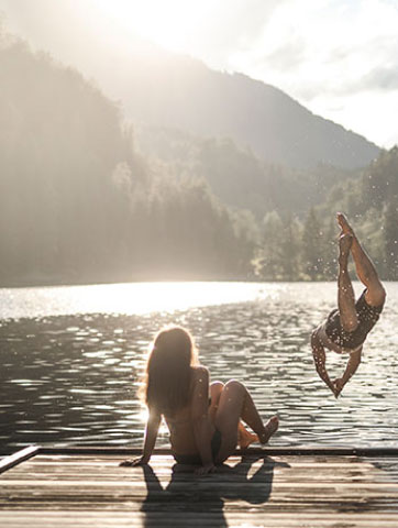 A person is sitting on a dock by a sunlit lake, watching another person mid-air performing a flip into the water. The scene is surrounded by misty mountains and bathed in warm sunlight.