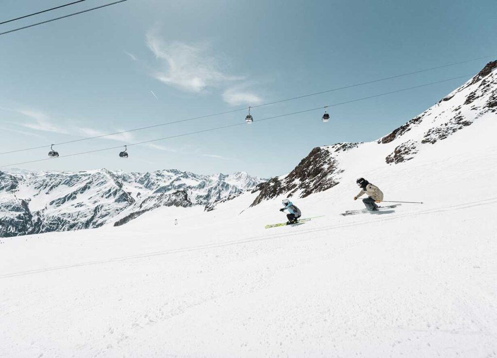 Zwei Skifahrer gleiten an einem klaren Tag einen schneebedeckten Hang hinunter, umgeben von hohen, schroffen Bergen. Der Himmel ist blau mit ein paar dünnen Wolken. Im Hintergrund ist eine Reihe von Skiliftgondeln zu sehen, die über der schneebedeckten Szene schweben.